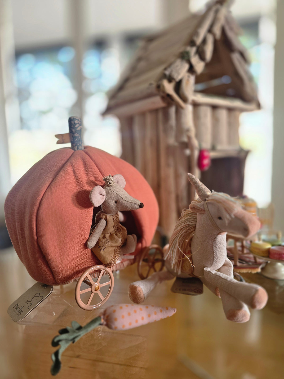 A soft fabric dollhouse scene featuring a small mouse in a pumpkin carriage next to a unicorn, with a tiny wooden house and miniature accessories in the background.