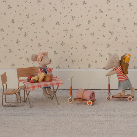 Two toy mice with a table and chairs, one on a scooter, against a floral wallpaper background.